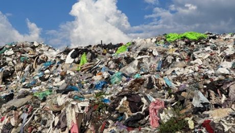 A large landfill displays a mountain of discarded textiles and mixed waste, with vibrant clothing colors contrasting against a blue sky and white clouds Garbage Dump